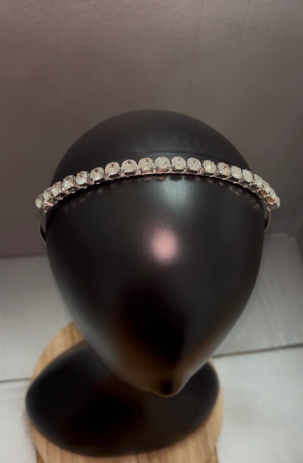 A silver-toned headband adorned with a single row of clear rhinestones, displayed on a black mannequin head bust.