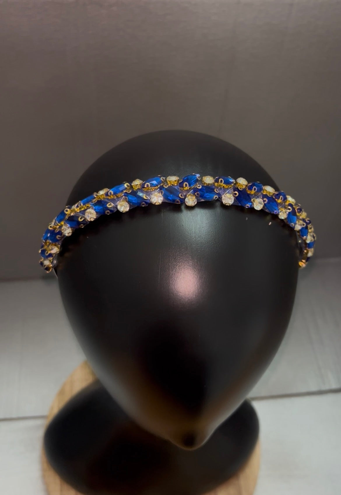 A gold-tone headband embellished with blue and clear rhinestones, displayed on a black mannequin head.