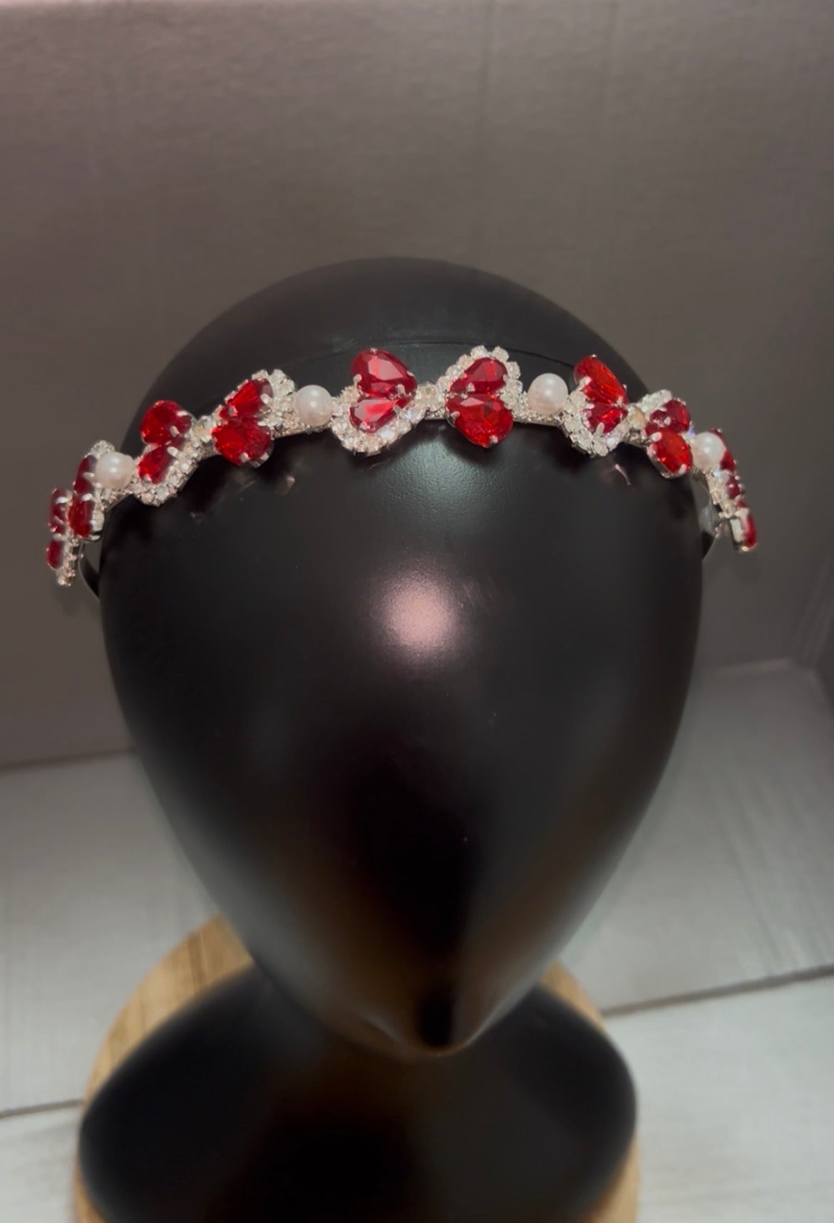 A silver tiara headband decorated with red heart-shaped crystals and white pearls, displayed on a black mannequin head.