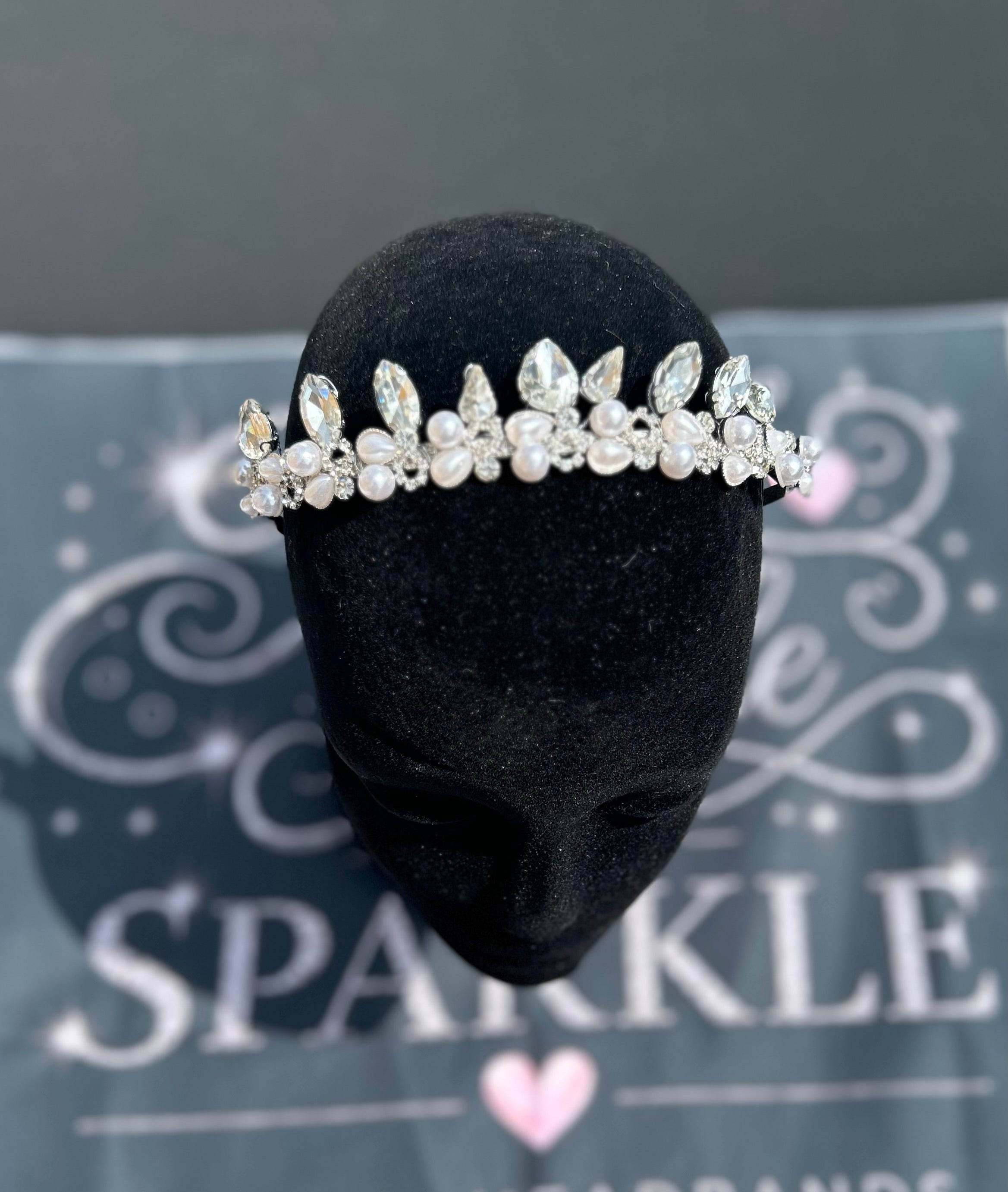A silver-toned floral tiara with clear rhinestones and white faux pearls, displayed on a black velvet mannequin head. The background shows a gray branded box with the word 'SPARKLE'.