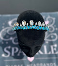 A sparkling headband with clear rhinestones and bright blue faceted crystals, displayed on a black velvet mannequin head. The background shows a branded gray card with the word 'SPARKLE' and 'THORAS HEADRANDES'.
